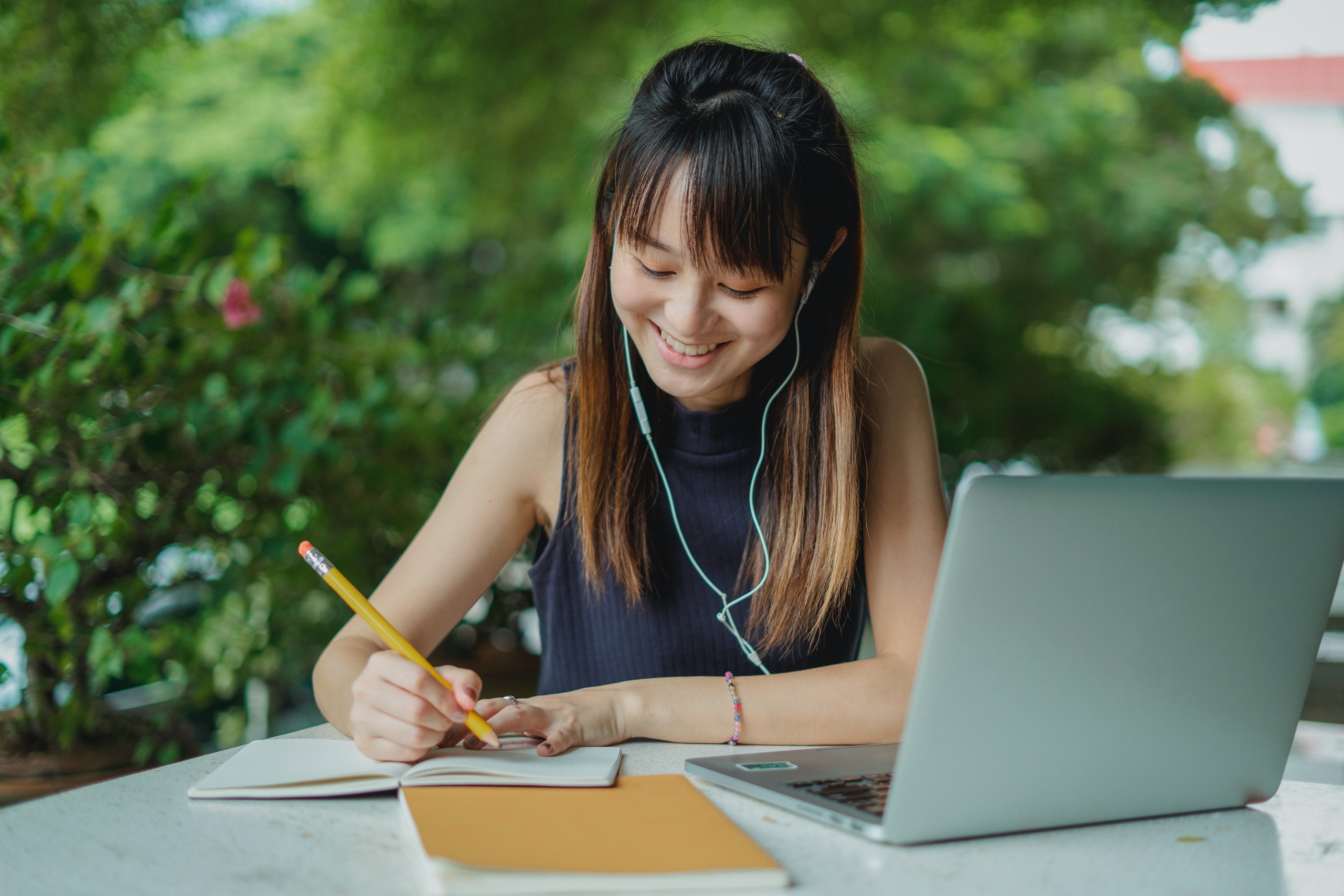 Alt=" A chinese lady seated and smiling while taking notes and writing down from a laptop as to a booklet"